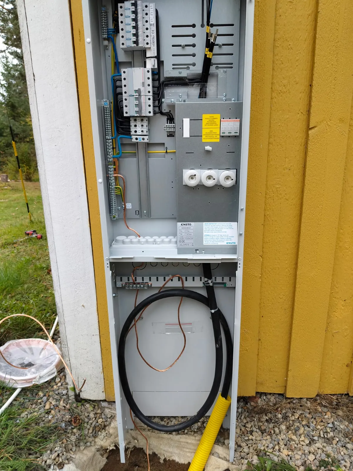 Avoin sähkötaulu kiinteistön ulkoseinällä, jossa näkyvissä sähkökaapelointeja ja sulakkeita.
